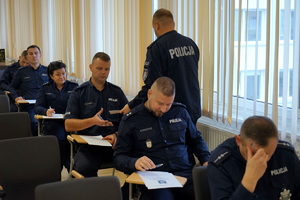 policjanci siedzą na krzesłach, słuchają organizatorów zawodów i przystępują do pisania testu