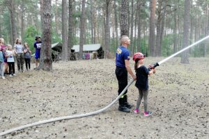 Harcerze symulują gaszenie pożaru wężem strażackim.