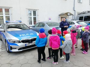 Dzieci słuchają prelekcji policjantki na dziedzińcu komendy