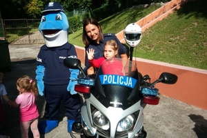Na motocyklu siedzi policjantka z dziewczynką, obok stoi duża maskotka