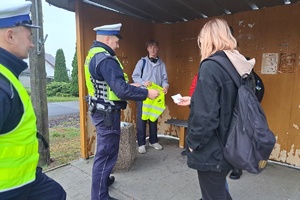 Policjanci drogówki wręczają kamizelki odblaskowe młodzieży stojącej na przystanku autobusowym