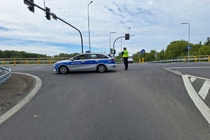 Policjant ruchu drogowego stoi na skrzyżowaniu przy radiowozie, zabezpiecza drogę