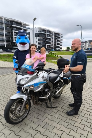 Policjant stoi przy policyjnym motocyklu, obok kobieta trzyma na ręku dziecko