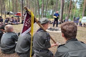 Harcerze siedzą na ziemi, słuchają policjanta