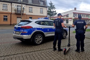 Policjanci trzymający hulajnogę, obok stoi radiowóz